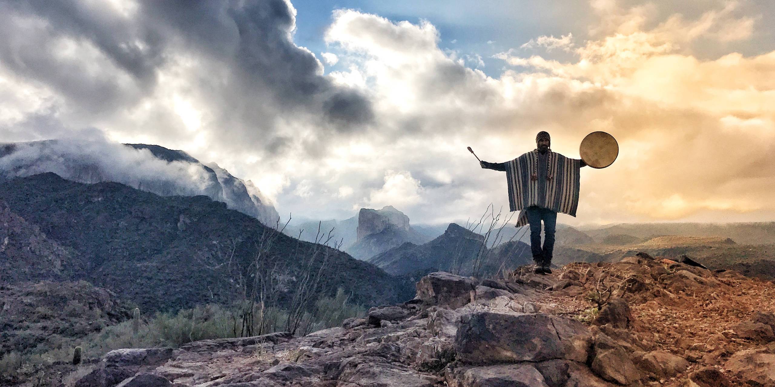 A person on a high ridge holds a round frame drum raised against an open sky.