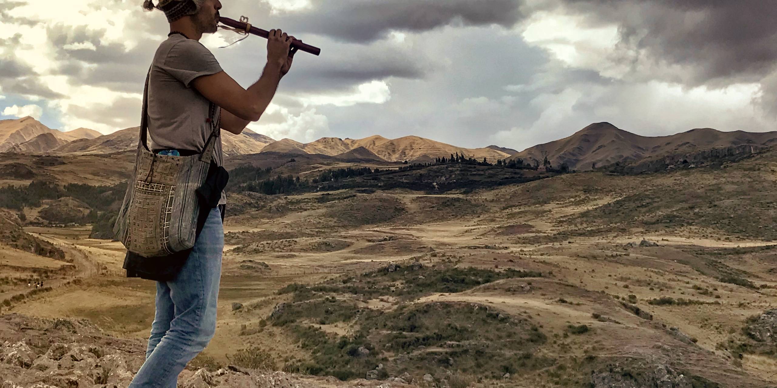 A person playing a wooden flute on a high ridge, the land falling away on both sides.