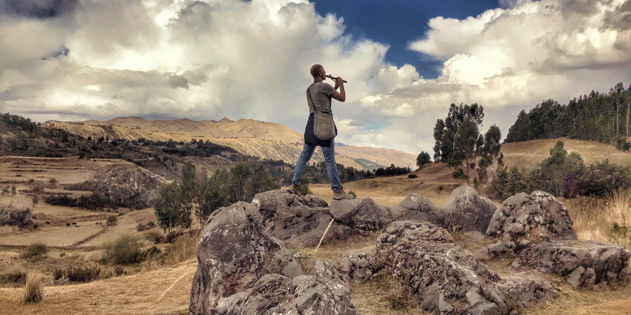 A person seated on high rocks playing a wooden flute, open country falling away below.