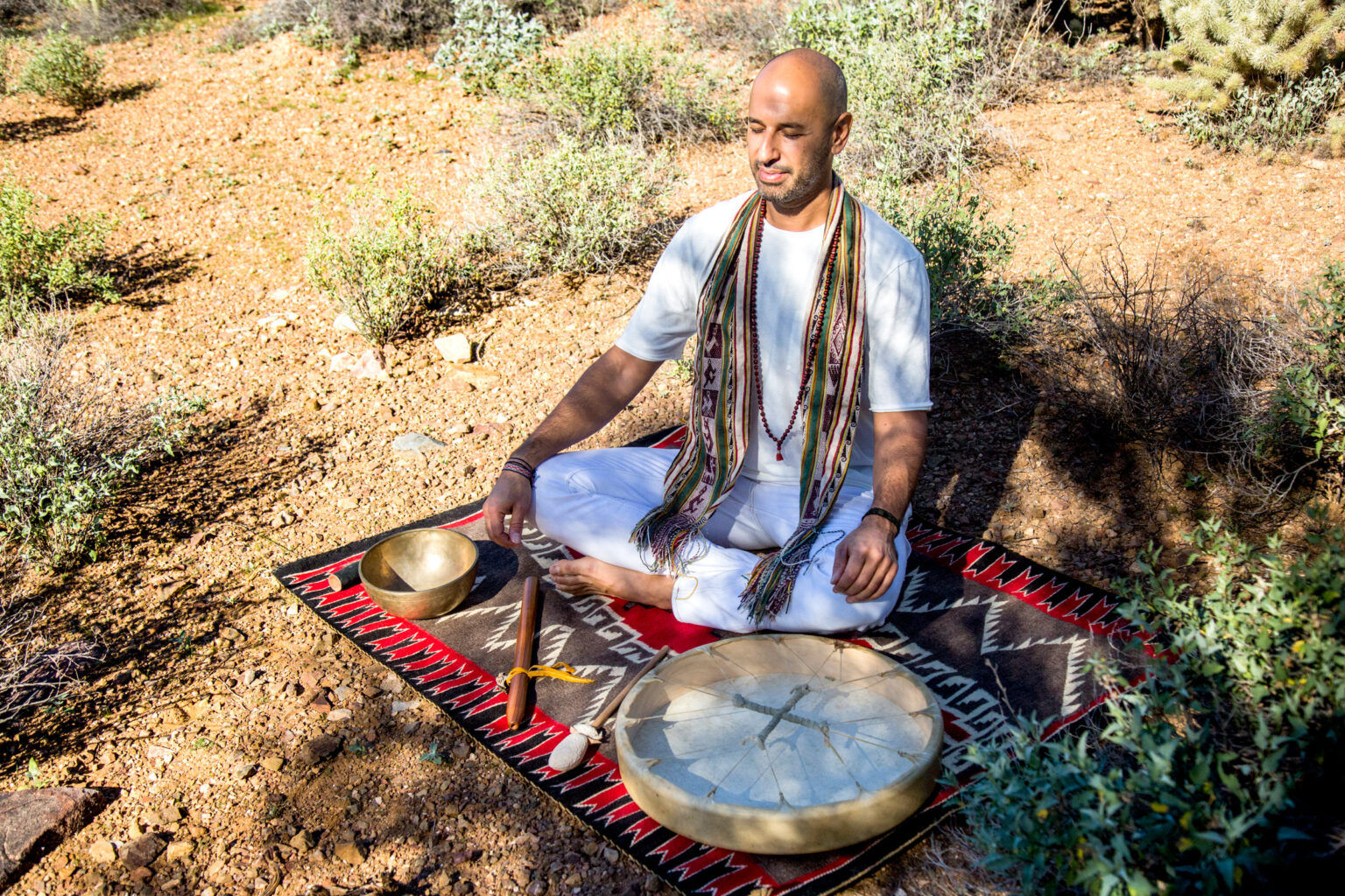 A man in white sits cross-legged on a patterned rug laid on the desert ground, with two bowls before him.